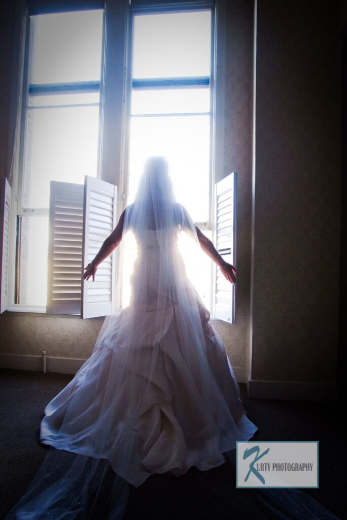 Bride standing in front of tall sunlit windows, wearing a layered couture gown and cathedral veil, symbolizing elegance and intentional wedding design.