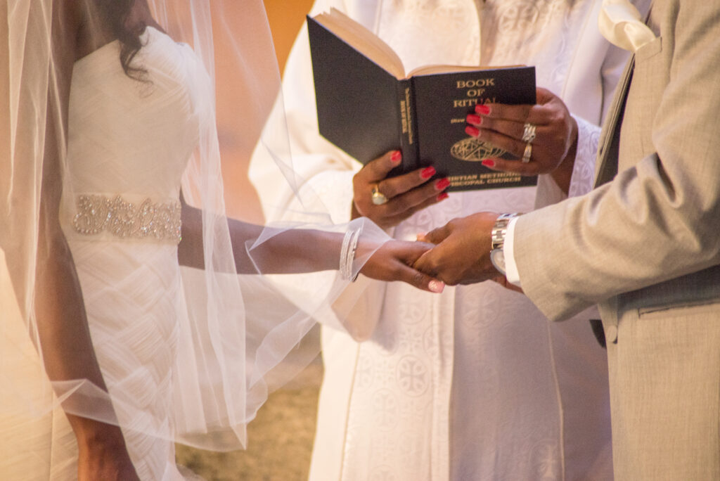 Close-up of a wedding ceremony moment where the bride and groom hold hands as the officiant reads from a book of rituals, highlighting a sacred and emotional exchange of vows planned by Marshecka Weddings.