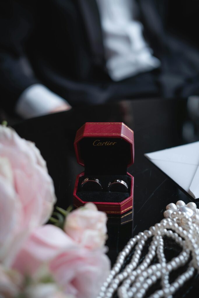 Luxury Cartier wedding rings in a red velvet box, surrounded by soft pink roses and pearl strands, styled elegantly on a black tabletop.
