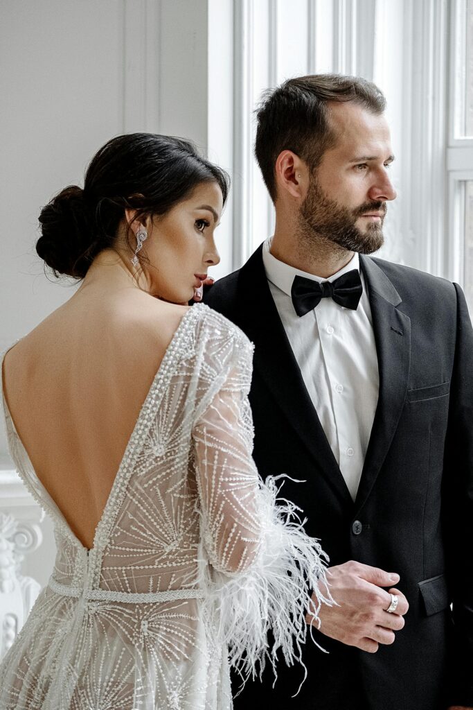 Bride in intricately beaded, feather-accented gown standing beside groom in classic black tuxedo — a timeless moment of modern garden opulence and editorial romance.