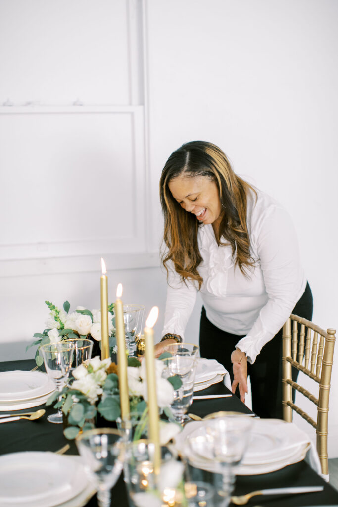 Marshecka, lead planner and designer, smiling warmly while refining a candlelit tablescape — a candid moment of joy and presence during event preparation
