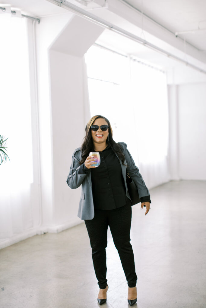 Marshecka Rodgers, luxury wedding and event planner, smiling in a chic gray blazer and black outfit while holding a celebratory glass in bright white studio. The photo captures confidence, elegance, and joy, reflecting her Fabulous Friday birthday reflections.