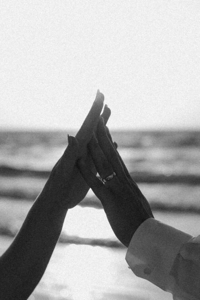Bride and groom’s hands touching in black and white at the ocean, capturing timeless love and the essence of a wedding celebration.