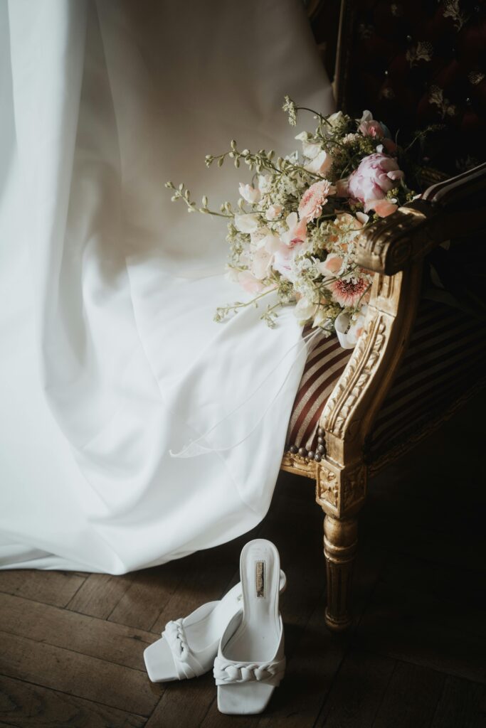 Elegant wedding detail with a cascading bridal bouquet of ivory florals resting on an ornate gold chair beside designer heels and a draped gown, a timeless, luxury-inspired vignette.