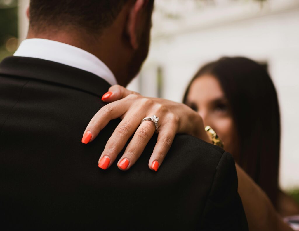 A bride rests her hand on her groom’s shoulder, her engagement ring catching the light as they share a quiet, intimate moment together.
